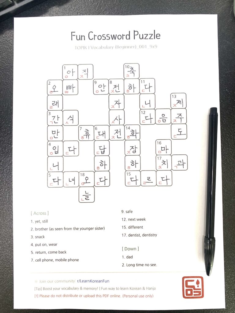 A printed Korean vocabulary crossword puzzle worksheet for TOPIK 1 beginners resting on a dark desk with a black pen. The page shows a 9x9 grid filled with Hangul words and English hints at the bottom.